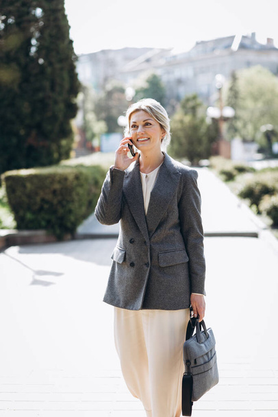 Mature blond smiling business woman walks the city streets and speaks on the phone - Foto, Imagen