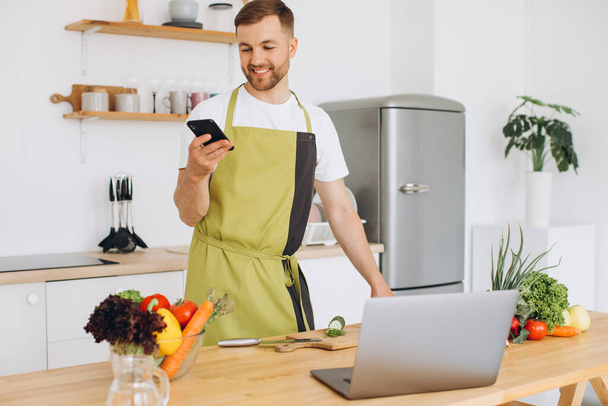 Attractive young man is cooking in the kitchen with a laptop on the table, talking on a smartphone. - Foto, afbeelding