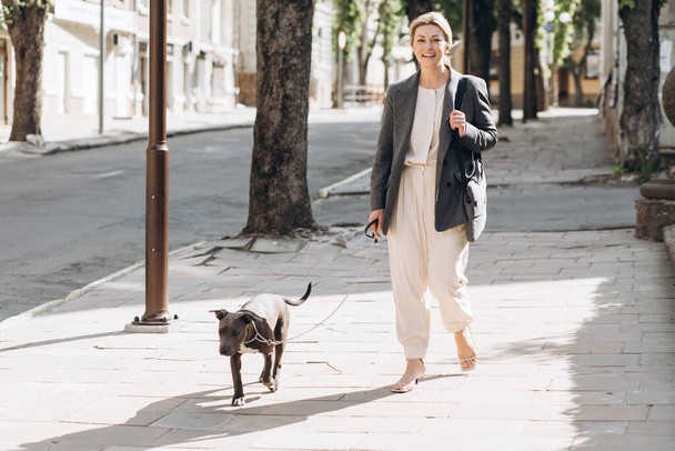 Madura rubia de negocios mujer en una chaqueta gris sonriendo paseando a su perro raza amstaff y hablando por teléfono - Foto, imagen