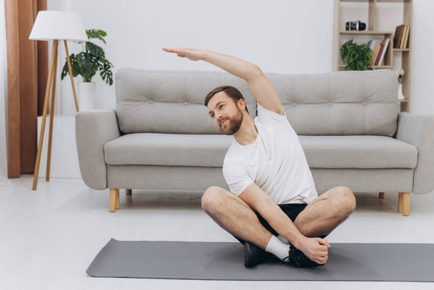 Active young cheerful man doing morning gymnastic at home, sitting on yoga mat and stretching, copy space - Foto, imagen