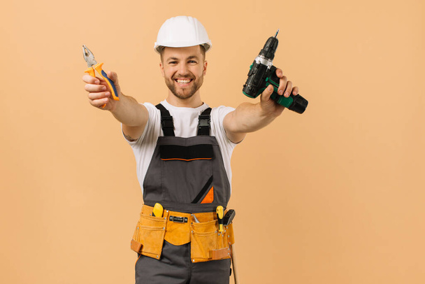 Позитивний чоловічий ремонтник в домашніх умовах показує викрутку і плоскогубці на бежевому фоні
 - Фото, зображення
