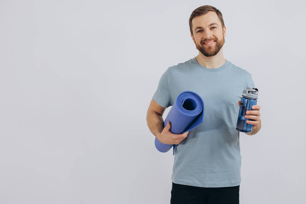 Portrait of handsome bearded man in sportswear holding yoga mat and bottle of fresh water on white background, copy space - Valokuva, kuva
