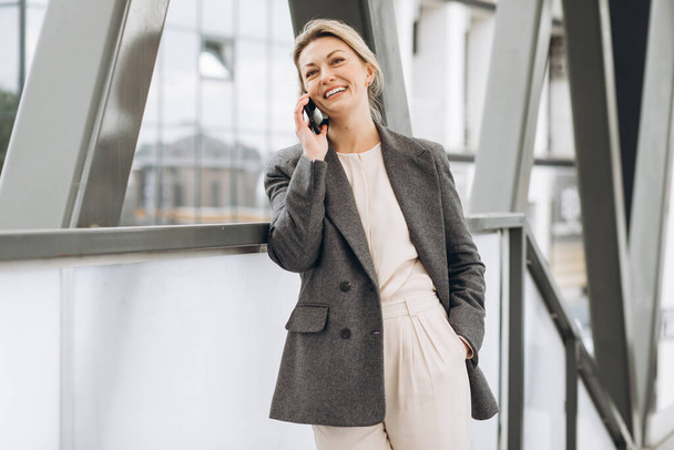 Portret pięknej dojrzałej kobiety biznesu w garniturze i szarej kurtce uśmiechającej się i rozmawiającej przez telefon na nowoczesnym miejskim tle - Zdjęcie, obraz