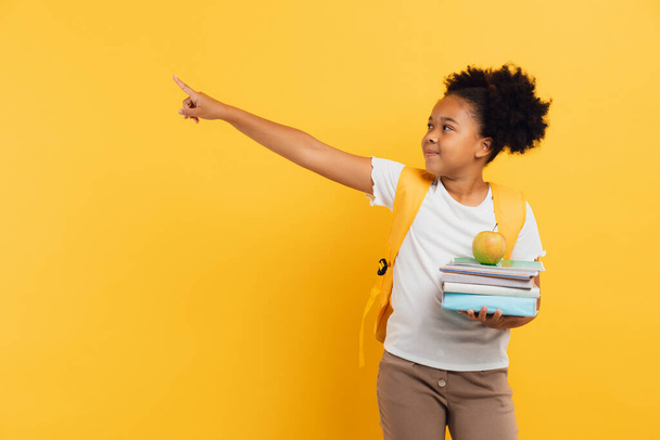 アフリカ系アメリカ人の女子学生がノートや本を持ち、黄色の背景に何かを表示し、スペースをコピー. - 写真・画像