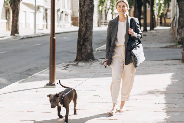 Mature blond business woman in a gray jacket smiling walking her dog breed amstaff and talking on the phone - Foto, afbeelding