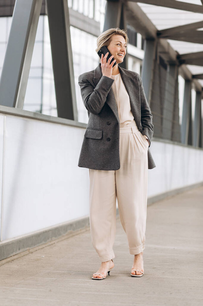 Retrato de una hermosa mujer de negocios madura en traje y chaqueta gris sonriendo y hablando por teléfono en un fondo moderno de edificios urbanos y de oficinas - Foto, imagen