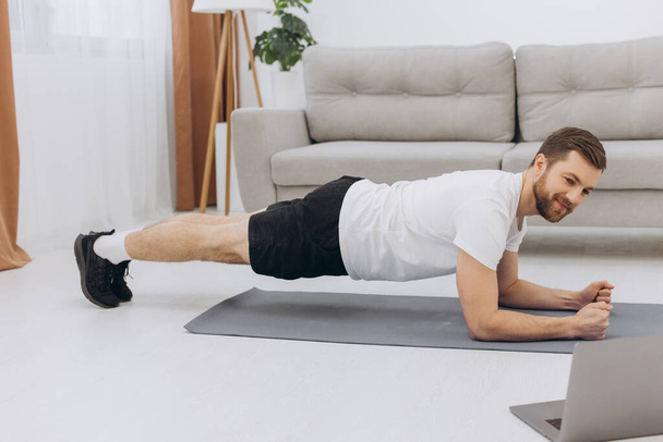 Otthon edzek. Sportos férfi csinál jóga deszka, miközben néz online bemutató laptopon, gyakorló a nappaliban, szabad hely - Fotó, kép