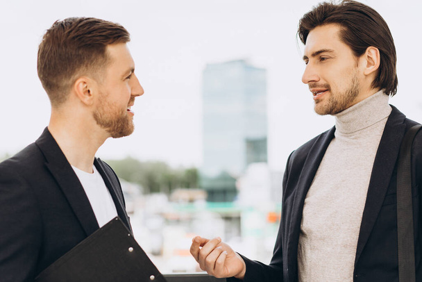 Twee moderne, vrolijke zakenlieden bespreken iets tegen de achtergrond van stedelijke kantoren en gebouwen - Foto, afbeelding
