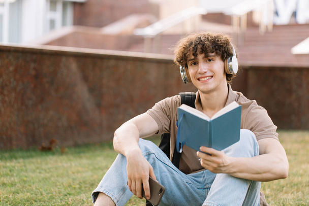 Atraktivní muž student ve sluchátkách sedí na trávě dělá domácí úkoly v blízkosti univerzity za slunečného dne - Fotografie, Obrázek