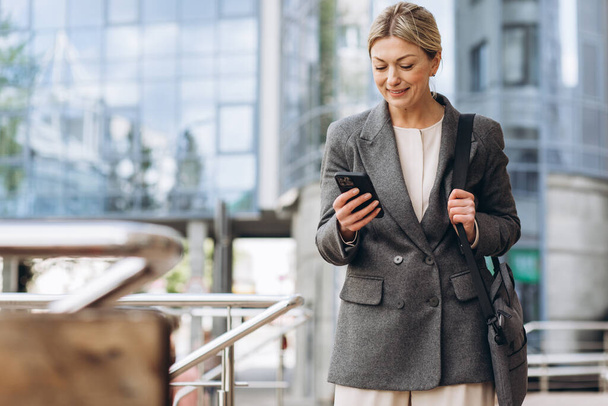 Portret van een mooie volwassen zakenvrouw in pak en grijze jas lachend en pratend aan de telefoon op de moderne stedelijke achtergrond - Foto, afbeelding