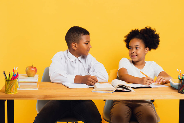 Χαρούμενη Αφροαμερικανή μαθήτρια και σχολιαρόπαιδο να κάθονται μαζί στο γραφείο και να μελετούν σε κίτρινο φόντο. Επιστροφή στο σχολείο έννοια. - Φωτογραφία, εικόνα