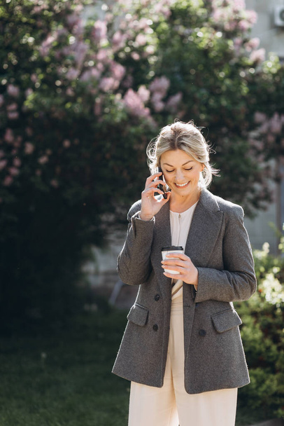 Starší blond usměvavá obchodní žena procházky v parku, pití kávy a mluvení po telefonu na jaře na pozadí šeříku květiny - Fotografie, Obrázek