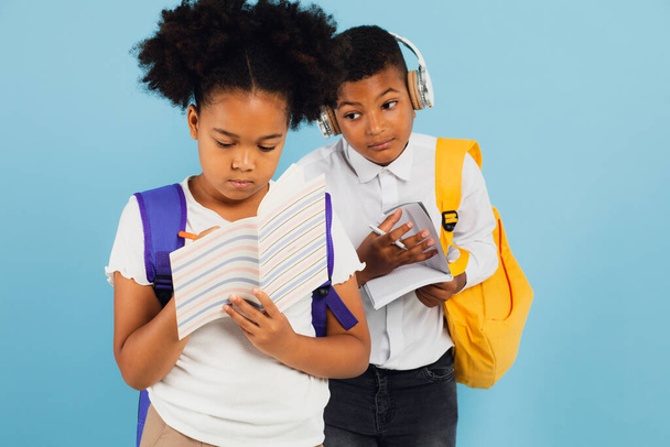 Mixed race schoolboy handing off homework to african american schoolgirl in school classroom on blue background, back to school concept. - Фото, изображение