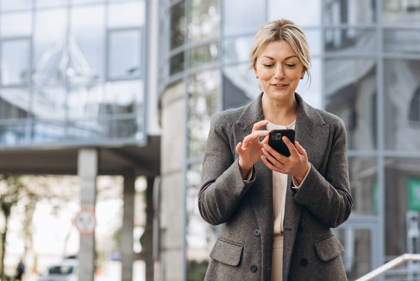 Portret van een mooie volwassen zakenvrouw in pak en grijze jas lachend en pratend op een telefoon op de moderne stedelijke en kantoorgebouwen achtergrond - Foto, afbeelding