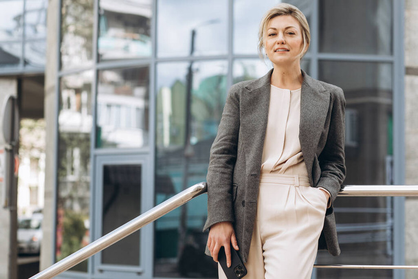 Retrato de uma bela mulher de negócios madura em terno e jaqueta cinza sorrindo e falando em um telefone no moderno urbano e edifícios de escritório fundo - Foto, Imagem