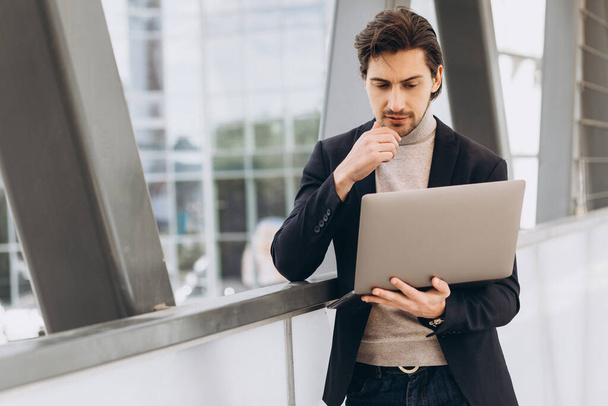 Portret van een knappe zakenman in pak met laptop op de achtergrond van stedelijke gebouwen en kantoren. - Foto, afbeelding