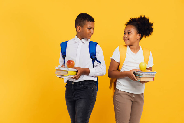 Gelukkig Afrikaans Amerikaans schoolmeisje en gemengde ras schooljongen met boeken naast elkaar op gele achtergrond. Terug naar schoolconcept. - Foto, afbeelding