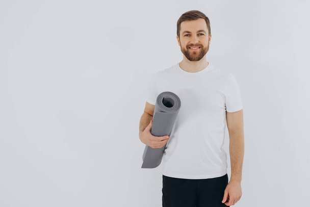 Портрет красивого бородатого мужчины в спортивной одежде с ковриком для йоги на белом фоне, пространство для копирования - Фото, изображение