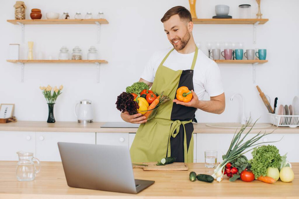 Portret van een gelukkige man thuis, man die groentesalade kookt kijkend naar de camera en lachend, groente snijdend, met laptop voor online kooktraining - Foto, afbeelding