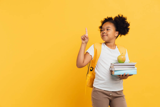 Colegiala afroamericana sosteniendo cuadernos y libros y mostrando algo sobre fondo amarillo, espacio para copiar. - Foto, Imagen