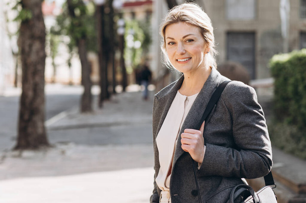 Portrait of a blond mature business woman smiling in a gray jacket on a background of the city - Foto, Imagem
