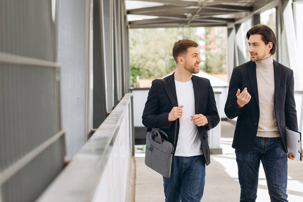 Zwei moderne, glückliche Geschäftsleute gehen über eine Stadtbrücke und diskutieren etwas vor dem Hintergrund städtischer Büros und Gebäude - Foto, Bild