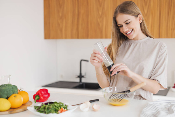 Feliz mujer rubia bonita en pijama está preparando el desayuno de huevos y sal entre verduras frescas en la cocina en casa. - Foto, Imagen