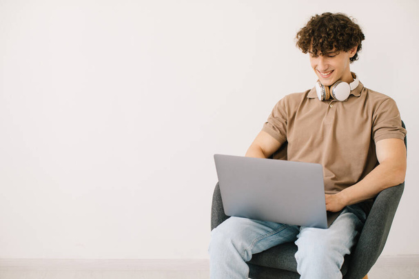 Attractive young man using laptop, wearing headphones while sitting in armchair against white wall, copy space. Millennial male communicating online, working or studying remotely on portable pc - Fotoğraf, Görsel