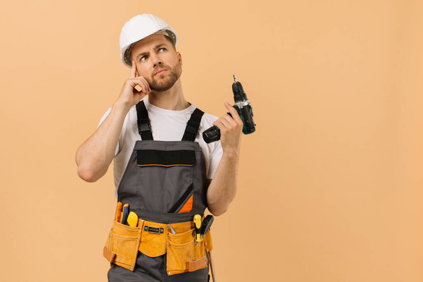 Positiver männlicher Reparateur zu Hause, der einen Schraubenzieher in der Hand hält und auf beigem Hintergrund denkt - Foto, Bild