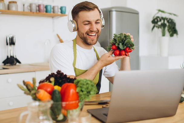 Happy man in headphones sitting at kitchen table and preparing salad, holding radish and showing it to laptop. - Фото, зображення
