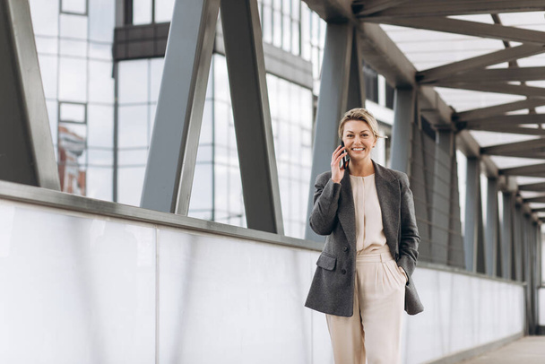Portret pięknej dojrzałej kobiety biznesu w garniturze i szarej kurtce uśmiechającej się i rozmawiającej przez telefon na nowoczesnym tle budynków miejskich i biurowych - Zdjęcie, obraz