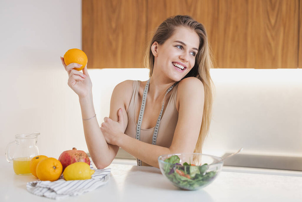 Happy attractive millennial woman in sportswear with measuring tape holding fresh orange for energy recovery after workout in kitchen - Fotografie, Obrázek