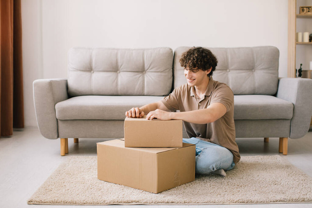 Happy young smiling curly man opening box with ordered goods gifts, presents at home on couch. Online shopper male customer opening online shop parcel. - Φωτογραφία, εικόνα