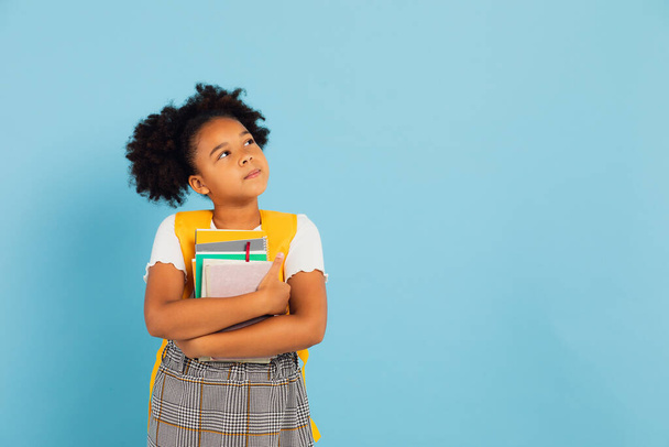 Счастливая афроамериканская школьница, держа в руках книги на синем фоне, возвращается в школу. - Фото, изображение