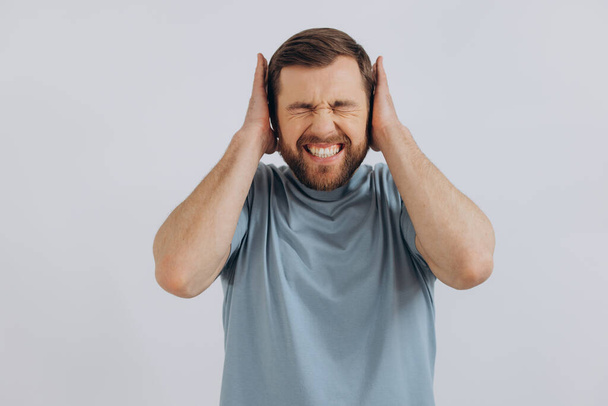 Retrato de un hombre barbudo moderno de mediana edad con una camiseta azul que muestra emociones cubriéndose las orejas sobre un fondo blanco - Foto, imagen