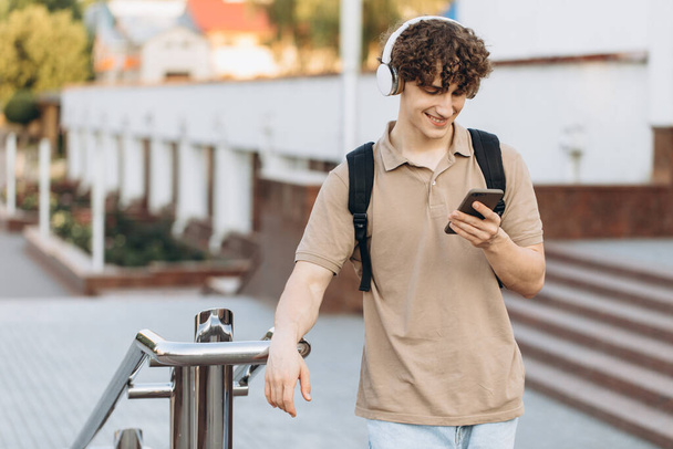Atraktivní kudrnatý mladý muž univerzitní nebo vysokoškolský student s telefonem procházky po areálu - Fotografie, Obrázek