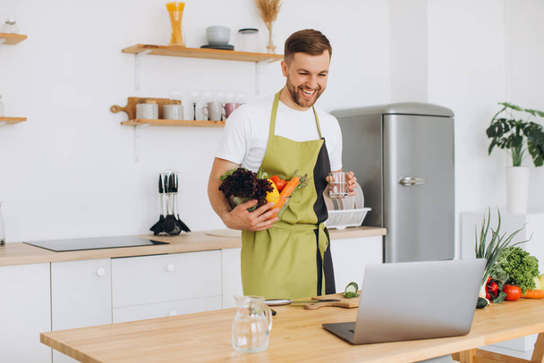 Πορτρέτο του ευτυχισμένος άνθρωπος στο σπίτι, άνθρωπος μαγείρεμα σαλάτα λαχανικών κοιτάζοντας κάμερα και χαμογελώντας, τεμαχίζοντας λαχανικά, χρησιμοποιώντας φορητό υπολογιστή για online προπόνηση μαγειρικής - Φωτογραφία, εικόνα