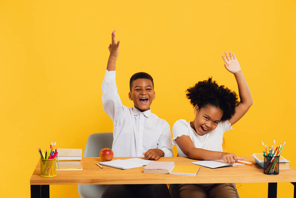 Happy african american female schoolgirl and schoolboy sitting together at desk and stretching arms up on yellow background. Back to school concept. - Photo, Image