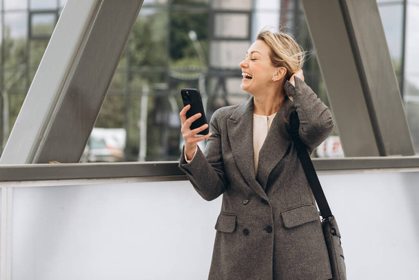 Porträt einer schönen reifen Geschäftsfrau im Anzug und grauer Jacke, die vor dem modernen urbanen Hintergrund lächelt und telefoniert - Foto, Bild