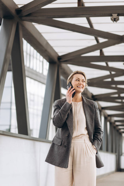 Portrait of a beautiful mature business woman in suit and gray jacket smiling and talking on the phone on a modern urban and office buildings background - Fotoğraf, Görsel