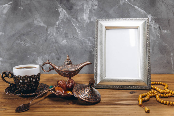 Composition for Ramadan. Silver frame with copy space or mockup, near traditional islamic tableware with dates and coffee on wooden table. - 写真・画像