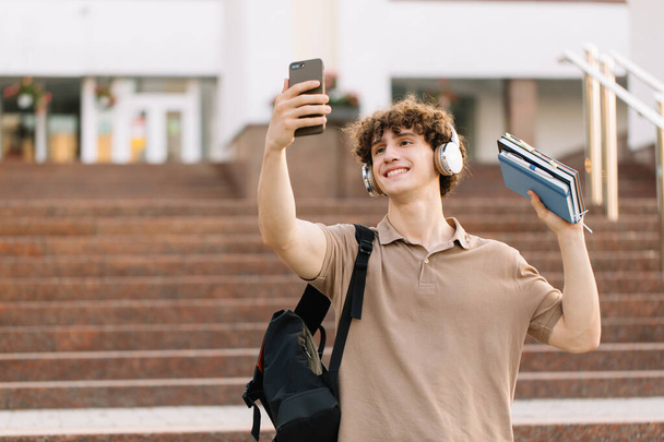 Çekici kıvırcık saçlı, kulaklıklı, gülümseyen ve kitaplarla selfie çeken erkek öğrencinin portresi sınavları geçti. - Fotoğraf, Görsel