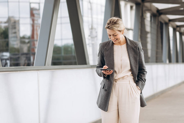 Portrait of a beautiful mature business woman in suit and gray jacket smiling and talking on the phone on a modern urban and office buildings background - Photo, Image