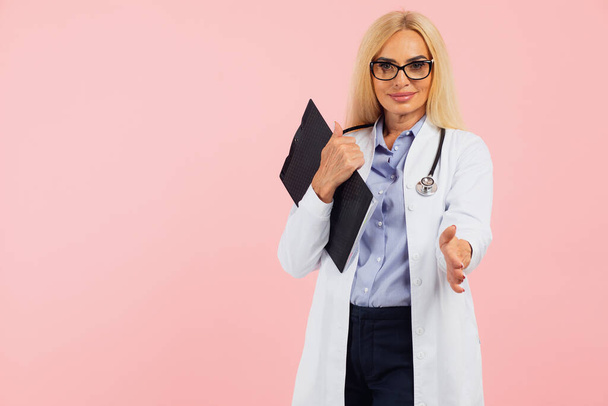 Médico mujer madura en gafas con estetoscopio celebración carpeta y dar respeto a la mano en el fondo rosa con espacio para copiar - Foto, imagen