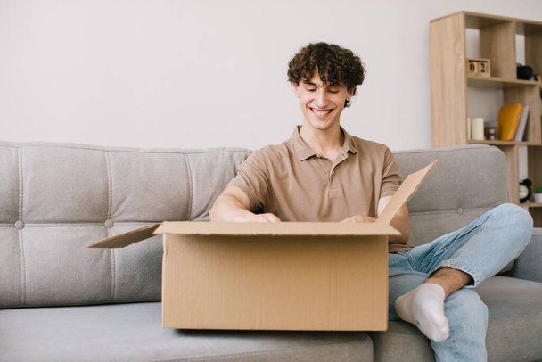 Happy young smiling curly man opening box with ordered goods gifts, presents at home on couch. Online shopper male customer opening online shop parcel. - Foto, Imagen