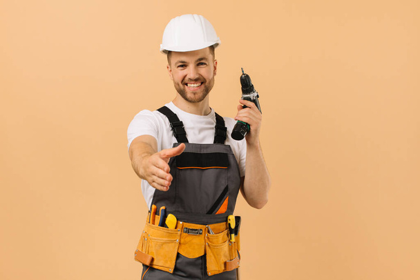 Reparador masculino positivo en casa sosteniendo un destornillador y extendiendo su mano para saludar sobre un fondo beige - Foto, imagen