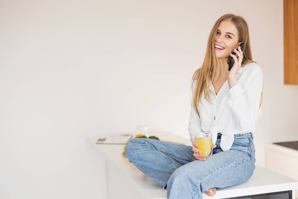 Mutlu ve gülümseyen sarışın kadın mutfak masasının üstünde oturmuş portakal suyu içiyor ve telefonda konuşuyor. - Fotoğraf, Görsel