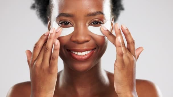 Face, skincare and happy black woman with eye mask in studio isolated on a gray background. Dermatology portrait, cosmetics and laughing female model apply facial collagen patches for healthy skin - Footage, Video
