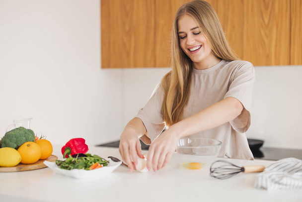 Onnellinen kaunis blondi nainen pyjamassa valmistaa aamiaista munista ja tuoreista vihanneksista keittiössä kotona.. - Valokuva, kuva