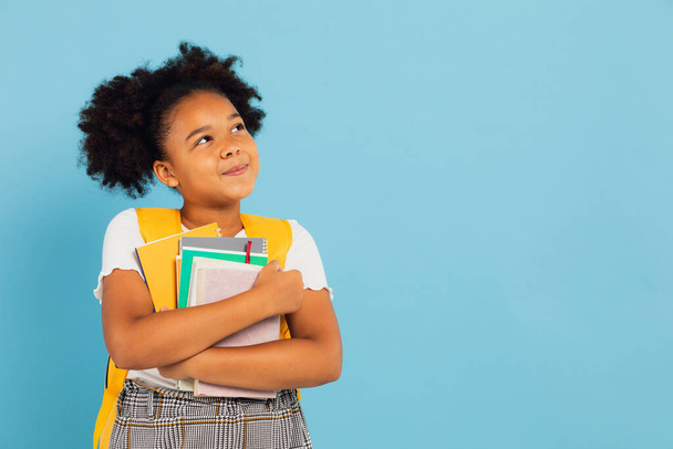 Happy African American schoolgirl holding books on blue background, back to school concept. - Photo, image